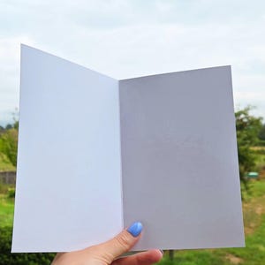 Pu&ograve; includere: Un biglietto di auguri aperto e vuoto con interno bianco. Il biglietto &egrave; tenuto contro uno sfondo di fogliame verde e un cielo nuvoloso. Il biglietto &egrave; pronto per un messaggio personalizzato.