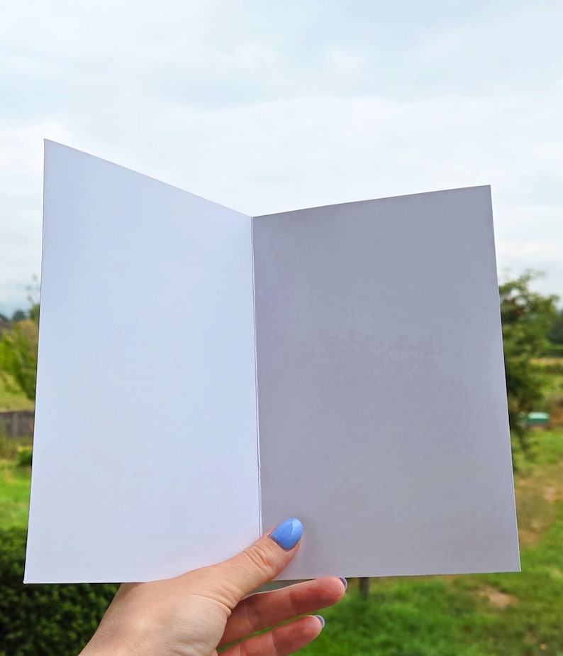 Pu&ograve; includere: Un biglietto di auguri aperto e vuoto con interno bianco. Il biglietto &egrave; tenuto contro uno sfondo di fogliame verde e un cielo nuvoloso. Il biglietto &egrave; pronto per un messaggio personalizzato.