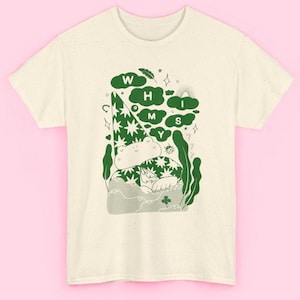 Può includere: T-shirt color crema con una stravagante illustrazione verde di una rana con un cappello a stelle. Il design include il testo "WHIMS" in lettere a forma di nuvola, insieme ad altri elementi decorativi. La maglietta è in materiale morbido.