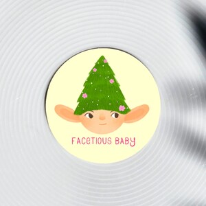 May include: A circular sticker featuring a cartoon illustration of a baby-like face wearing a green Christmas tree hat with pink flowers. The text "FACETIOUS BABY" is written in pink below the face.