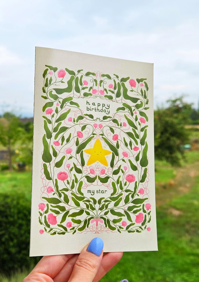 Pu&ograve; includere: Biglietto di auguri con un design floreale e le parole "buon compleanno" e "my star". Il biglietto presenta una stella gialla al centro, circondata da fiori rosa e foglie verdi. Lo sfondo &egrave; color crema chiaro.