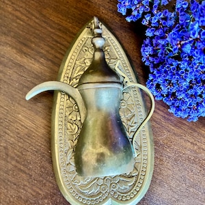 May include: A vintage brass coffee pot with a curved spout and handle, mounted on an ornate, leaf-shaped base. The base features intricate floral patterns. The pot is a warm, antique gold color, and the base has a slightly darker patina. A cluster of blue flowers is in the background.