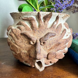 May include: A ceramic planter featuring a textured, off-white base with a prominent, raised brown relief of a sun-like face. The face has stylized rays, defined eyebrows, and a protruding, open-mouthed spout at the bottom center. The surface has a speckled, earthy finish with visible clay texture throughout the exterior.