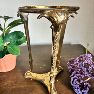 May include: A vintage brass plant stand with a circular top and three legs. The legs are adorned with ram's head details and decorative patterns. The stand is designed to hold a small potted plant, and is placed on a wooden surface.