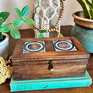May include: A rectangular dark brown wooden box featuring two square decorative tiles inlaid on the lid. The tiles display a vibrant circular pattern in yellow, blue, and red. The box includes a small metal latch on the front for closure.