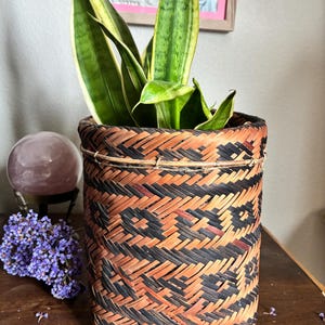 May include: A woven basket planter with a geometric pattern in black and brown, holding a snake plant with green and yellow striped leaves. The basket is cylindrical and sits on a wooden surface. A purple flower arrangement and a pink crystal ball are also visible.
