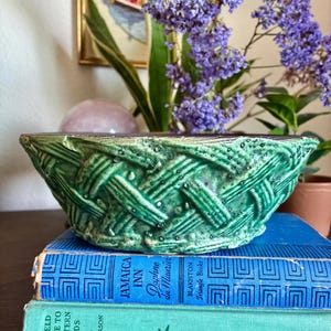 May include: A green ceramic bowl with a textured, woven basket-weave pattern on the exterior. The bowl is resting on top of two stacked hardcover books, one blue titled Jamaica Inn and one light green titled A Field Guide to Western Birds.