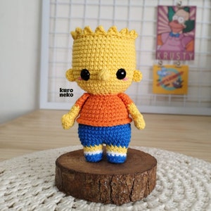 May include: A crocheted plush toy of Bart Simpson from The Simpsons. The toy is yellow with a blue shirt, orange shirt, and white shoes. The toy has black eyes and a black nose.