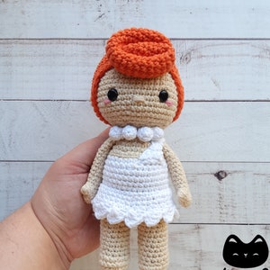 Puede incluir: Una muñeca de crochet con pelo naranja, un vestido blanco y un collar de perlas. La muñeca está de pie sobre una mano.