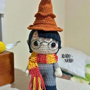 Pode incluir: Uma boneca de crochê com cabelo preto, óculos pretos redondos e uma cicatriz em forma de raio na testa. A boneca usa um chapéu pontudo laranja, um cachecol listrado de vermelho e amarelo com franjas e um conjunto cinza. Texto KURO NEKO.