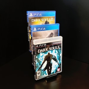 Puede incluir: Tres cajas de videojuegos apiladas una encima de la otra. La caja superior es azul y dice "PS4 DARK SOULS III". La caja del medio es azul y dice "PS4 DARK SOULS II". La caja inferior es negra y dice "PS3 DARK SOULS".
