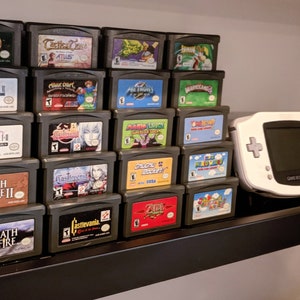 May include: A collection of video game cartridges for the Game Boy Advance, displayed on a shelf. The cartridges are arranged in rows, with the titles facing forward. The titles include "Golden Sun", "Final Fantasy", "Advance Wars", "Harvest Moon", "Breath of Fire", and "Gunstar Heroes".