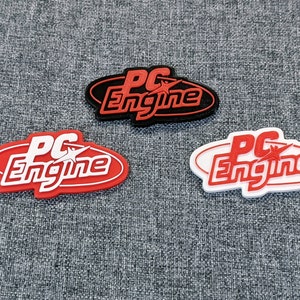 May include: Three PC Engine logos, one red, one white and one black. The logos are oval-shaped with a white star and the text "PC Engine" in a stylised font.