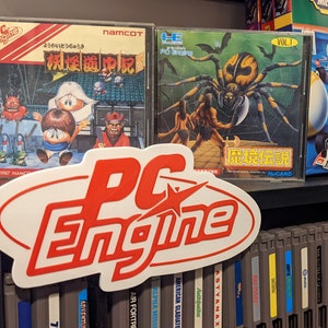 Könnte beinhalten: Eine Sammlung von Vintage-PC-Engine-Videospielkassetten, darunter "Youkai Dou no Yuuki" und "Majou Densetsu Gaiden", die in einem Regal mit einem rot-weißen PC-Engine-Logo ausgestellt sind.
