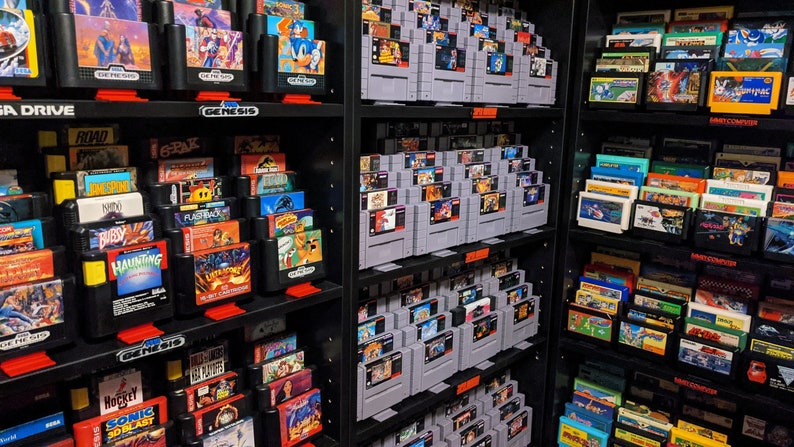 May include: A collection of vintage video game cartridges organized on shelves. The cartridges are for Sega Genesis, Super Nintendo, and Nintendo Entertainment System consoles. The cartridges are arranged by color and title.