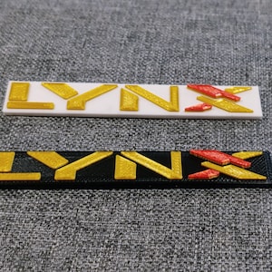 May include: Two versions of the word "LYNX" in gold lettering. The top version is on a white background with a red and gold "X" design. The bottom version is on a black background with a red and gold "X" design.
