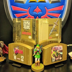 May include: A collection of gold-colored Nintendo video game cartridges, including The Legend of Zelda, Zelda II: The Adventure of Link, and Ocarina of Time. The cartridges are displayed on a shelf with a gold-colored Triforce logo behind them. There are also figurines of Link and Ganondorf.