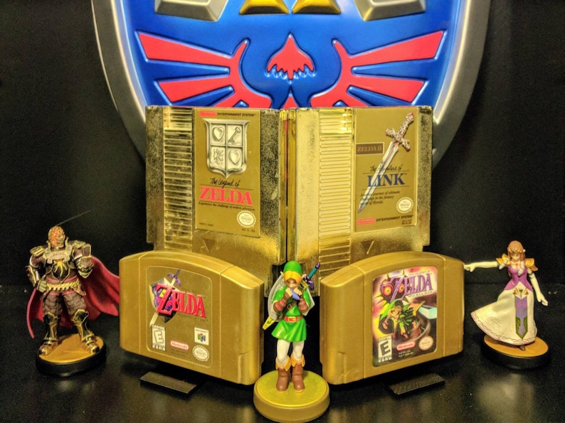 May include: A collection of gold-colored Nintendo video game cartridges, including The Legend of Zelda, Zelda II: The Adventure of Link, and Ocarina of Time. The cartridges are displayed on a shelf with a gold-colored Triforce logo behind them. There are also figurines of Link and Ganondorf.