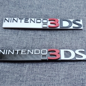 May include: Two black and white plastic Nintendo 3DS logos. The top logo is on a white background and the bottom logo is on a black background. The "3" in the logo is red.