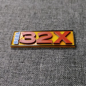 Peut inclure: Un panneau rectangulaire jaune et noir avec le texte "SEGA 32X" en lettres rouges en relief.