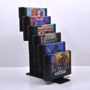 May include: A black plastic stand holds six Sega Genesis game cartridges. The cartridges are arranged vertically, with the game titles facing forward. The titles include "Sonic the Hedgehog", "Aladdin", "Street Fighter II", "Ecco the Dolphin", and "Mortal Kombat II".
