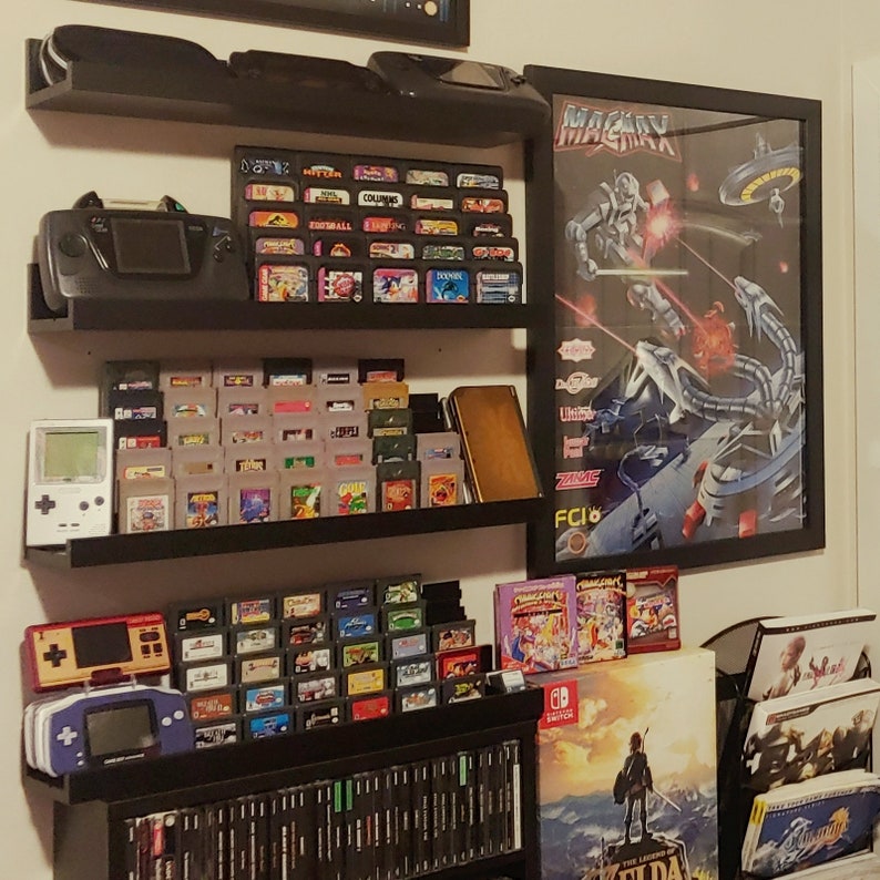 May include: A black shelf unit with multiple levels displaying a collection of video game cartridges, consoles, and posters. The top shelf features a Sega Genesis console and a poster for the game 'Maxx' with a futuristic robot design. The middle shelf displays a variety of game cartridges, including a Game Boy Advance and a Game Boy Color. The bottom shelf features a collection of video game cartridges and a Nintendo Switch game case.