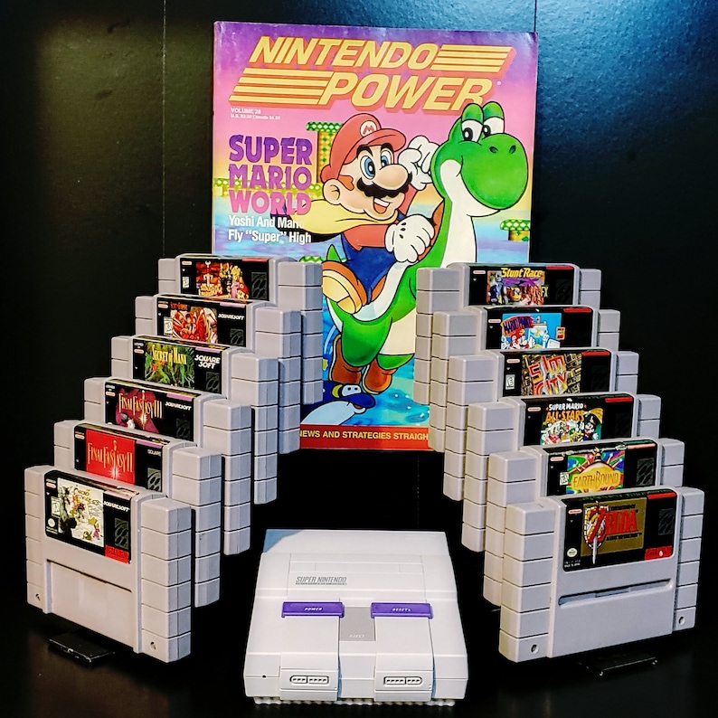 K&ouml;nnte beinhalten: Eine Sammlung von Super Nintendo Entertainment System (SNES) Spielkassetten, die in einem Regal ausgestellt sind. Die Kassetten sind in Reihen angeordnet und beinhalten Titel wie "Super Mario World", "Yoshi's Island" und "Stunt Race FX". Das Regal ist aus grauem Kunststoff und hat eine schwarze Basis. Im Hintergrund ist ein Exemplar des Nintendo Power Magazins mit Super Mario World zu sehen.