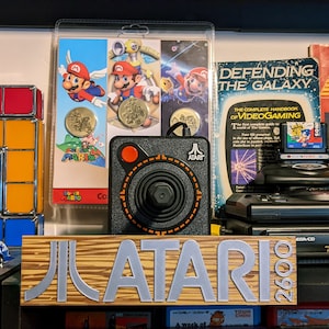 Può includere: Una console Atari 2600 vintage con una targa in legno che recita "Atari 2600". La console è su uno scaffale con altri oggetti per videogiochi, tra cui un joystick, una cartuccia di gioco e un libro sui videogiochi.