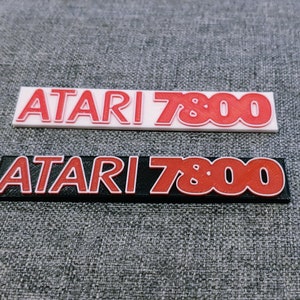 May include: Two versions of a black and white Atari 7800 logo. The top logo is white with red lettering. The bottom logo is black with red lettering.