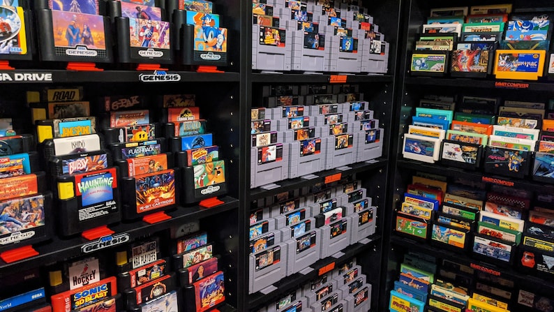 K&ouml;nnte beinhalten: Eine Sammlung von Vintage-Videospielkassetten, die in Regalen sortiert sind. Die Kassetten sind nach System geordnet, darunter Sega Genesis, Super Nintendo und Game Boy. Die Regale sind schwarz und die Kassetten sind in verschiedenen Farben.