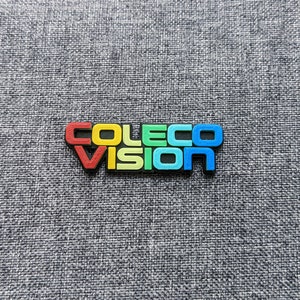 Puede incluir: Primer plano de un logotipo de plástico de colores para ColecoVision, una consola de videojuegos vintage. Las letras son rojas, amarillas, verdes, azules y negras.