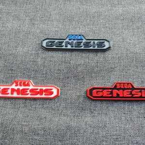 Op de afbeelding: Drie SEGA Genesis logo-emblemen in verschillende kleurenschema's: zwart en grijs, rood en wit en rood en zwart. De logo's zijn rechthoekig met de tekst SEGA Genesis in een vet lettertype.