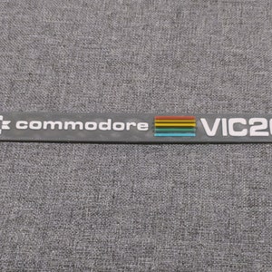 Może przedstawiać: Czarno-białe logo Commodore VIC20 z paskiem tęczy.