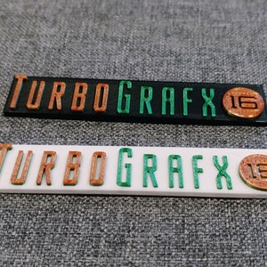 May include: Two close-up images of a black and white plastic plaque with the text "TURBOGRAFX 16" in orange and green letters.