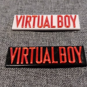 以下が含まれることがあります： 「VIRTUAL BOY」の文字が赤色で立体的にプリントされた長方形のネームプレートが2つ。1つは白、もう1つは黒。どちらも太字のサンセリフ体。レトロゲーム愛好家向けのアイテムです。