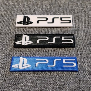 Puede incluir: Tres llaveros rectangulares de plástico con el logotipo de PlayStation y el texto "PS5" en blanco. Los llaveros son blancos, negros y azules.