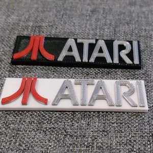 May include: Two Atari logo plaques, one with a black background and silver lettering, and the other with a white background and silver lettering. The Atari logo features a red stylized "A" with a red "J" on the left.