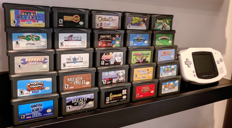 May include: A collection of video game cartridges for the Game Boy Advance, displayed on a shelf. The cartridges are arranged in rows, with the titles facing forward. The titles include "Golden Sun", "Final Fantasy", "Advance Wars", "Harvest Moon", "Breath of Fire", and "Gunstar Heroes".