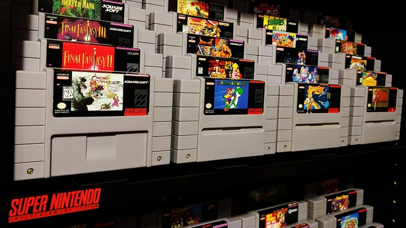 May include: A collection of Super Nintendo Entertainment System video game cartridges, displayed on a shelf. The cartridges are arranged in rows, with the labels facing forward. The cartridges are all in their original cases, and many of the labels feature colorful artwork.