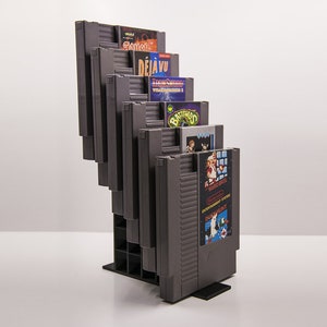 May include: A gray plastic stand with slots to hold multiple NES game cartridges. The cartridges are stacked vertically and feature various game titles, including "Deja Vu", "Batman", and "Duck Hunt".