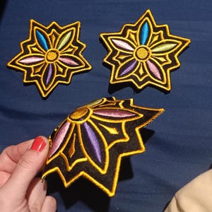 Könnte beinhalten: Gestickte Blumenaufnäher mit schwarzem Hintergrund und goldfarbenem Rand. Die Blütenblätter sind in Blau-, Grün-, Rosa- und Lilatönen gehalten, mit einem gelben Zentrum. Die Aufnäher sind sternförmig.