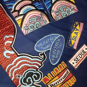 May include: Embroidered patches in various shapes and colors on a navy blue background. Designs include stylized waves, text that says "SLAY", "SEOUL", "Rrrrrrum!", and "HUNTRIX World Tour".
