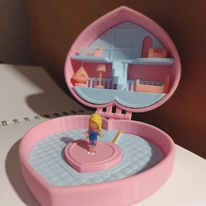 May include: A pink heart-shaped toy playset with a miniature doll. The playset opens to reveal a two-story interior with furniture. The doll has blonde hair and is wearing a blue outfit. The playset is on a white surface.