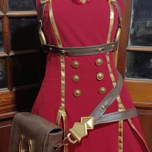 May include: A sleeveless red dress featuring a white collar with a red bow and a gold diamond-shaped emblem. The dress includes gold trim, six gold buttons, a brown belt with a gold buckle, and a brown side bag adorned with a gold horseshoe emblem.
