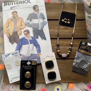 May include: A wooden surface displays a Butterick pattern, buttons, a necklace, and jewelry findings. The pattern features illustrations of jackets. Buttons are white and gold-toned. The necklace has colorful beads. A black square holds gold-toned earrings.