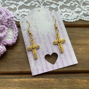 May include: Gold-colored cross earrings hanging on a lavender and white striped tag with a heart-shaped cutout. A crocheted lavender and white flower is on the left. The background is a wooden surface.