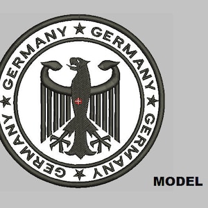 Puede incluir: Un parche bordado en blanco y negro con el águila alemana y la palabra "GERMANY" repetida alrededor del borde.