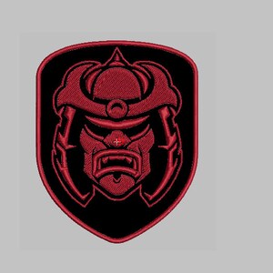 Peut inclure: Un écusson brodé représentant un visage de samouraï stylisé avec une expression féroce, en rouge et noir.