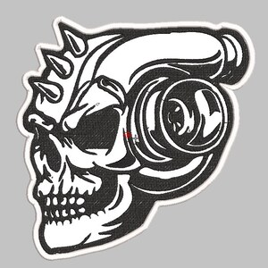 May include: Black and white embroidered patch of a skull with a turbocharger attached to the side of its head. The skull has a menacing expression and sharp teeth. The turbocharger has a detailed design with a large, round impeller.