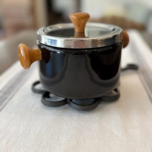 May include: A black pot with a shiny finish, featuring a silver metal lid and light brown wooden handles on the sides and top. The pot rests on a dark, circular, flower-shaped metal trivet base, providing a stable foundation for the cooking vessel.
